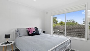 Un dormitorio con un ventanal grande, una cama con ropa de cama a rayas, una almohada rosa y una mesita de noche pequeña con una lámpara.