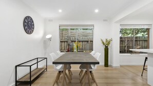 A modern dining area with a long table, white chairs, a clock on the wall, and a view of the outdoors through large windows. - Puebla Casa 17  Walk to Beach & Shops (TORQUAY)
