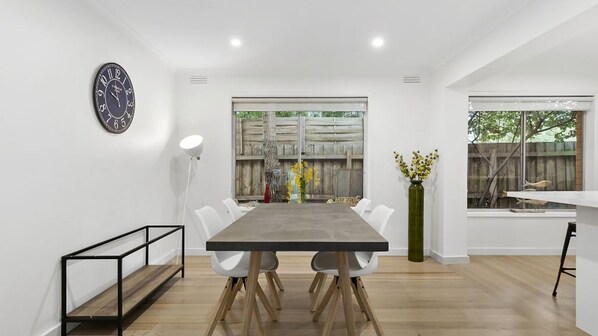 A modern dining area with a long table, white chairs, a clock on the wall, and a view of the outdoors through large windows. - Puebla Casa 17 Walk to Beach & Shops (TORQUAY)