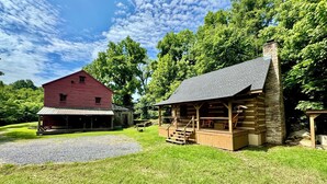 Une cabane rustique en rondins de bois, dotée d’une cheminée en pierre, d’une grange rouge et d’un porche abrité.