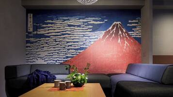 Habitación con un mural tradicional japonés del Monte Fuji, un sofá, una mesa de centro con tazas y una planta, y una manta oscura sobre el sofá.