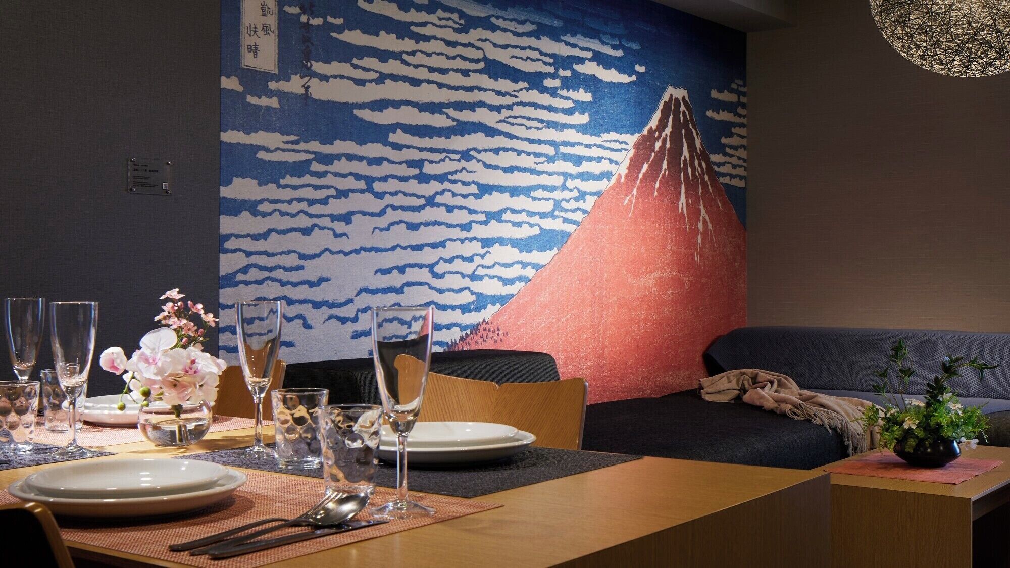 Un coin repas avec une table dressée pour deux personnes, des verres, des assiettes et un vase de fleurs. Une grande fresque représentant une montagne et des nuages orne le mur derrière la table.