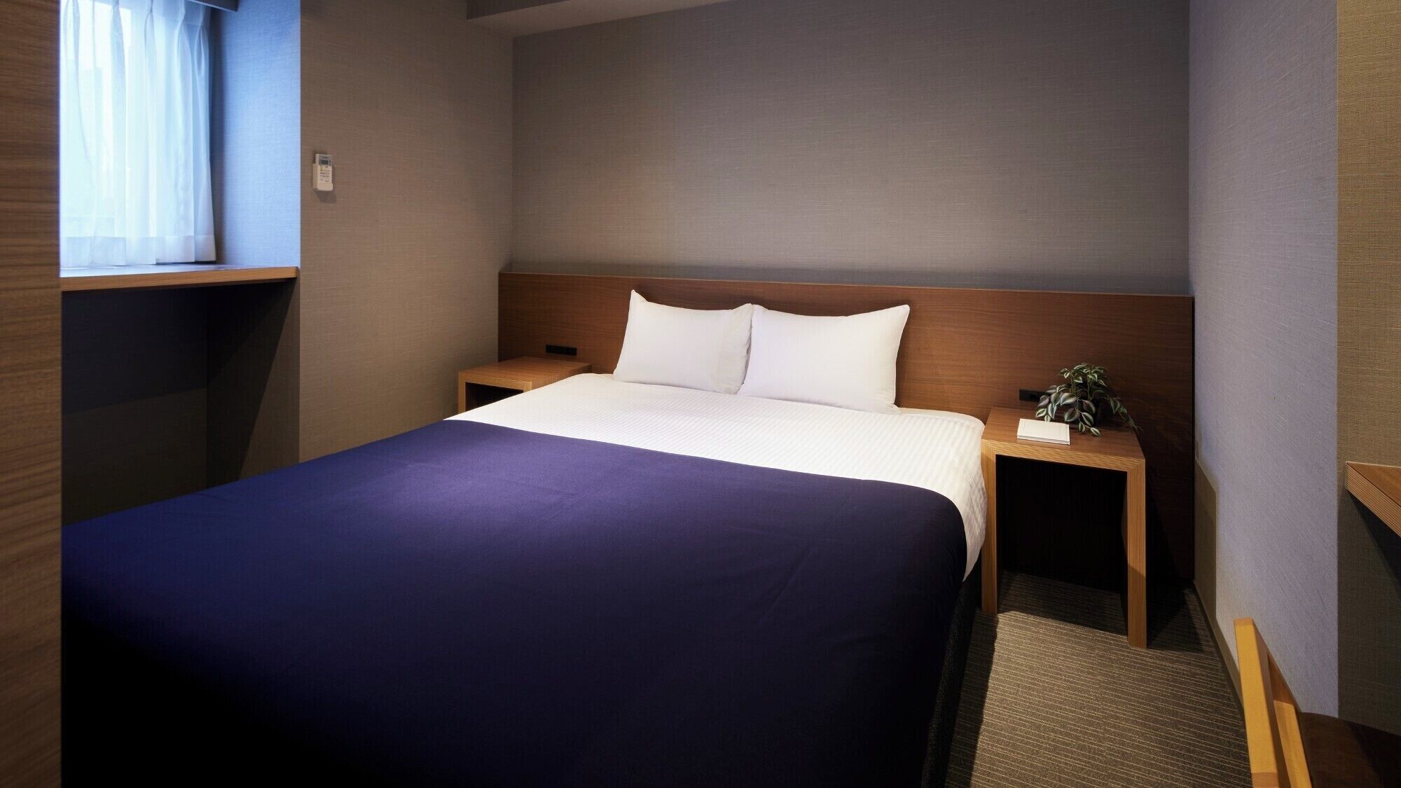 Una habitación de hotel con una cama grande, cabecera de madera, mesita de noche con una planta y una ventana con cortinas.