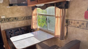 A compact kitchenette with a small table and bench, a window with curtains, and a wall-mounted sign. - Eagle- Camper at Sunset Bay  (Onanole)