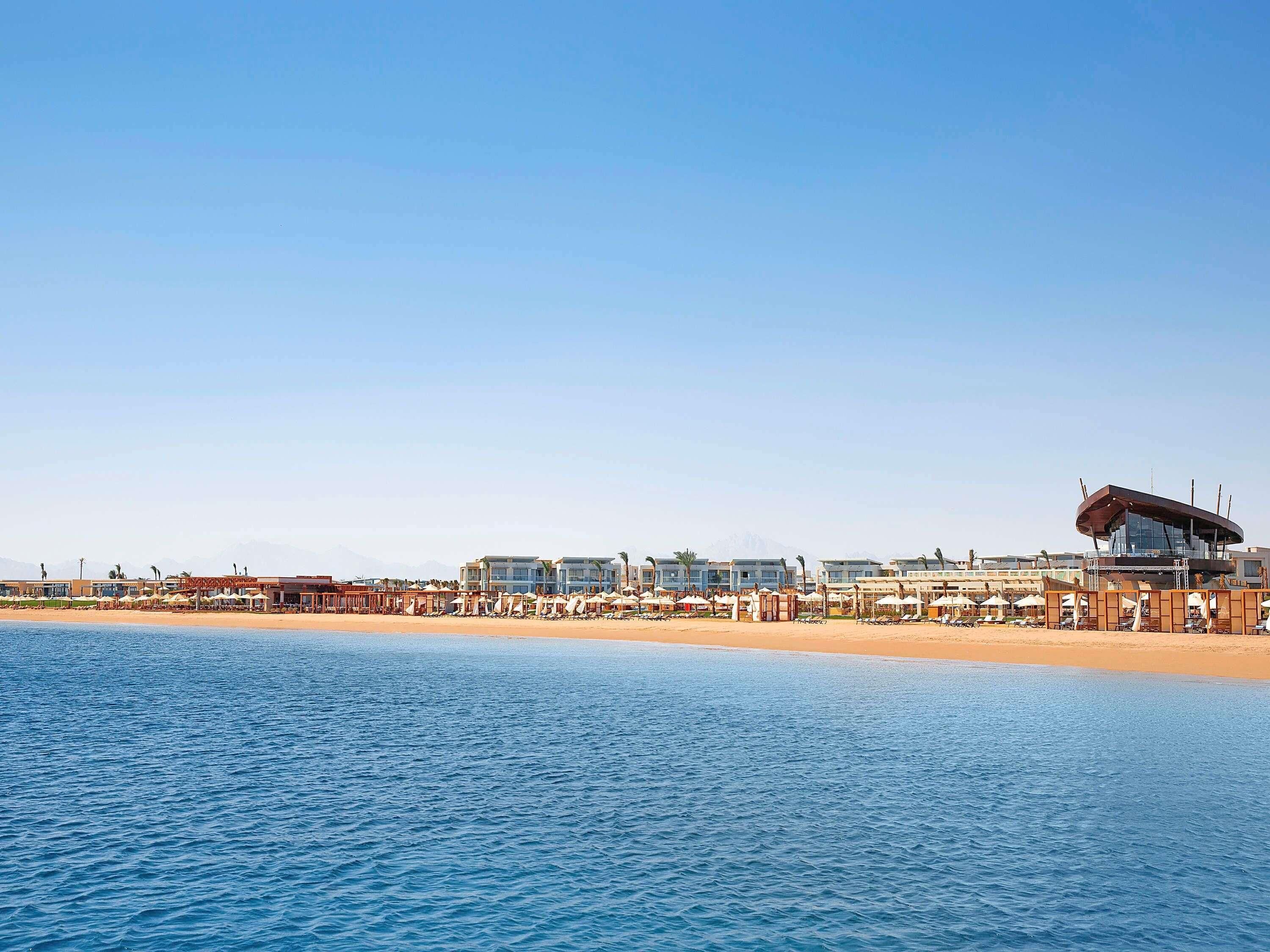 Un hôtel situé sur la côte, avec des chambres donnant sur la plage, une piscine et un grand bâtiment doté d’une structure de toit originale.