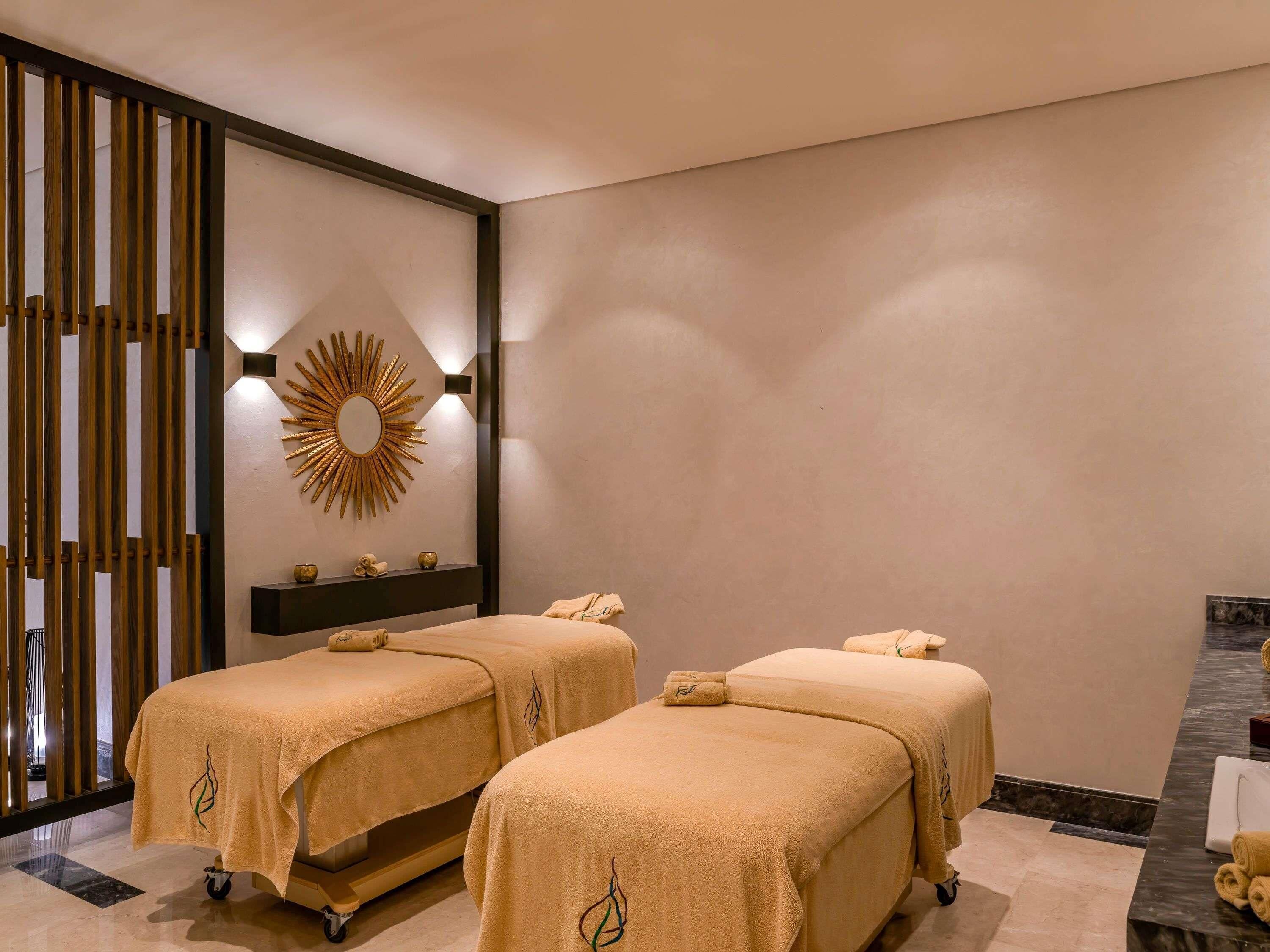 A spa room with two massage tables, a sunburst mirror, a wooden partition, and a marble floor.
