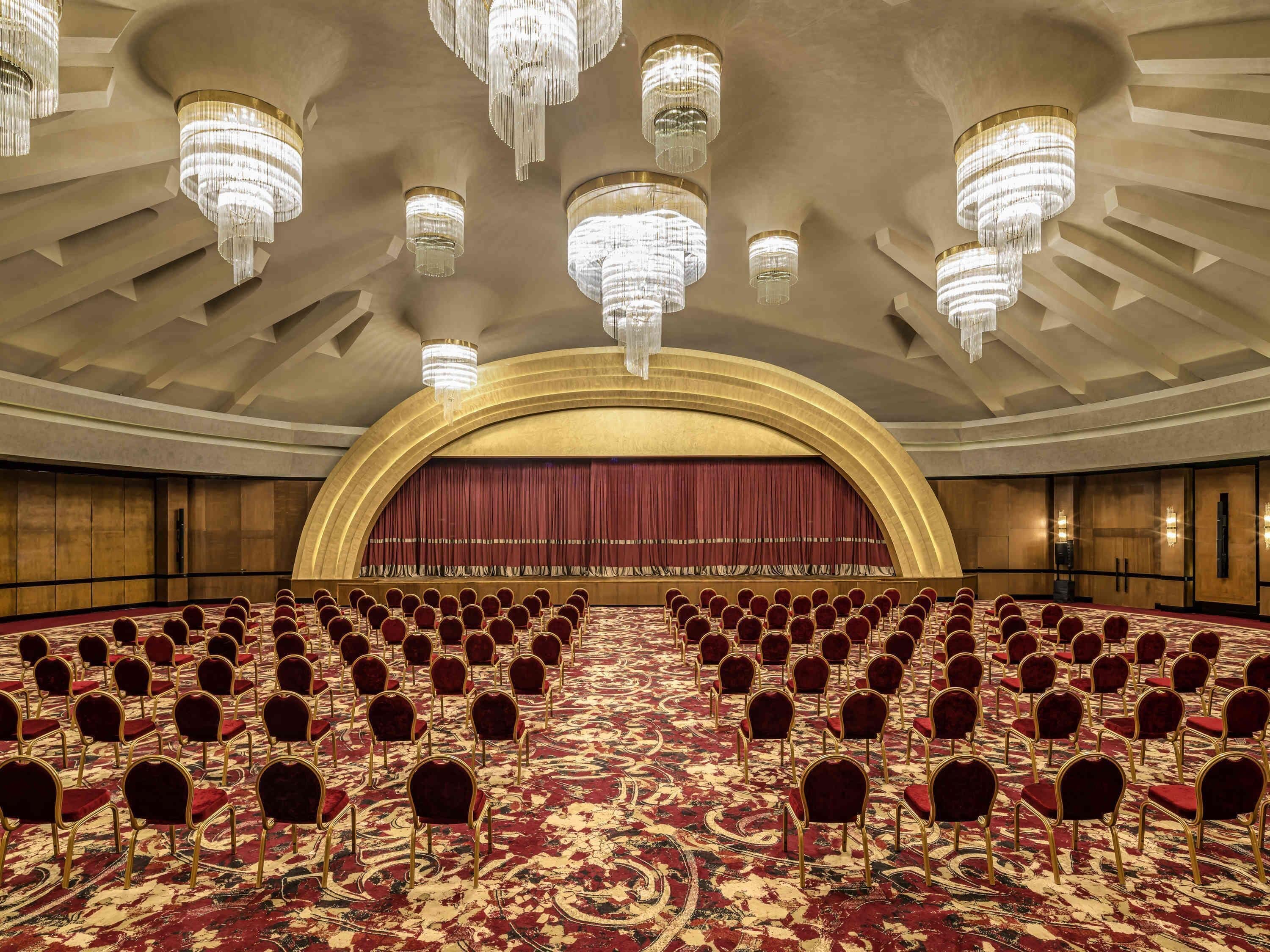 A large conference room with rows of chairs, a stage with a red curtain, and multiple chandeliers.