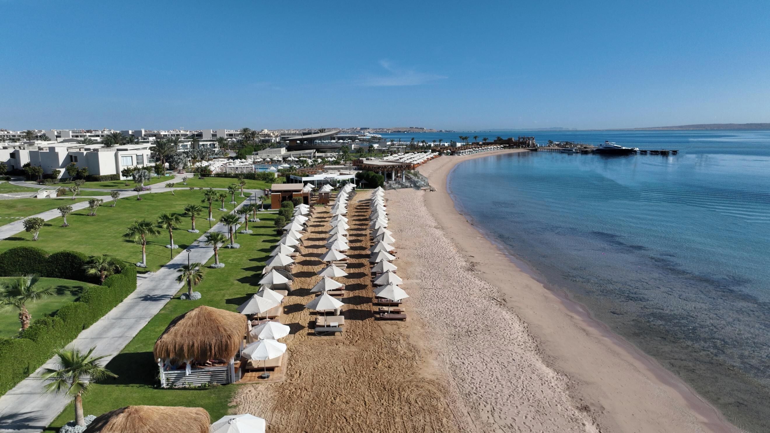 A beach resort with rows of sun loungers and umbrellas, a clear blue sea, and a distant view of a cityscape.