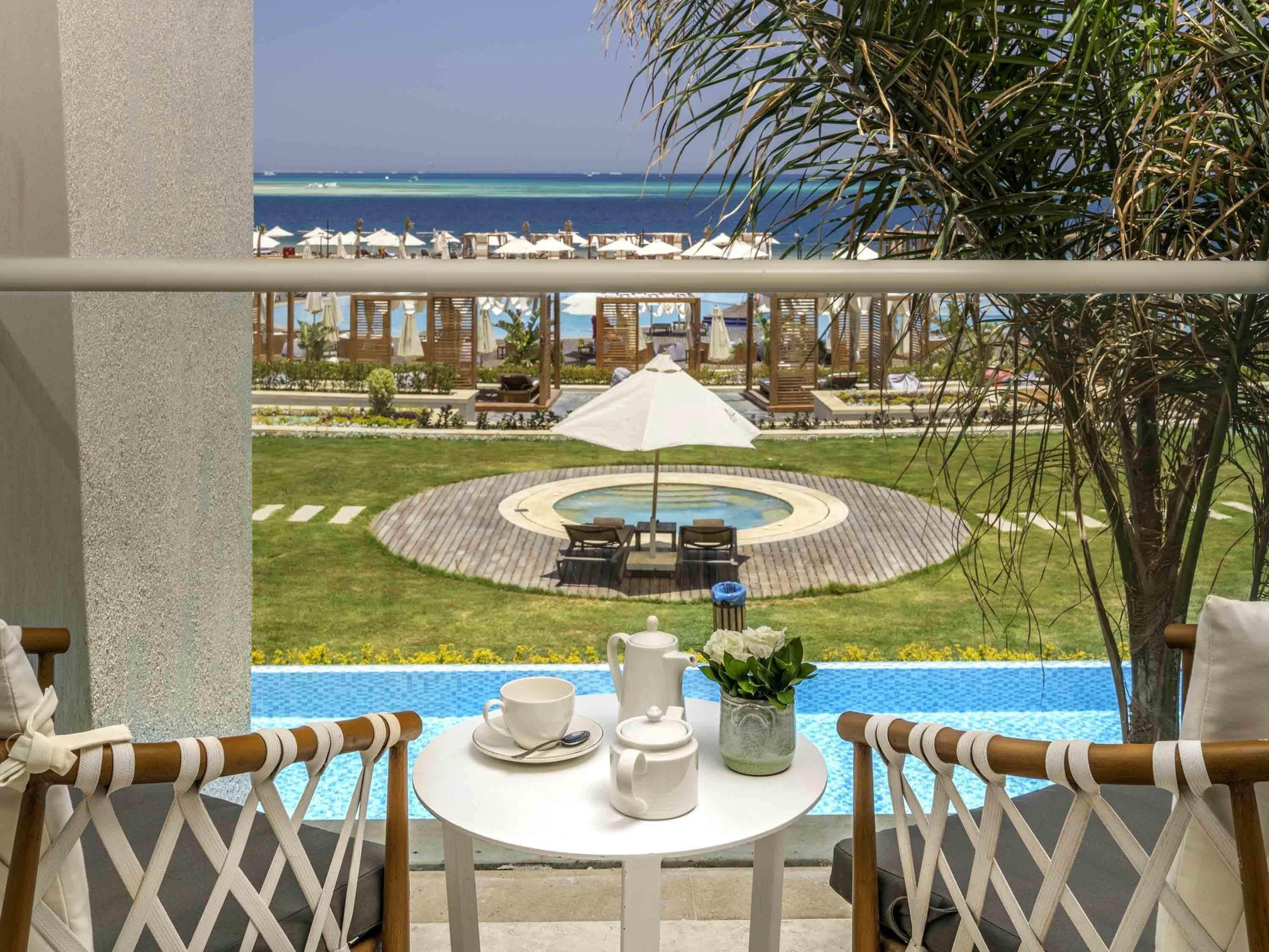 A balcony with a view of a pool, lounge chairs, and a dining table with a pitcher and cup.