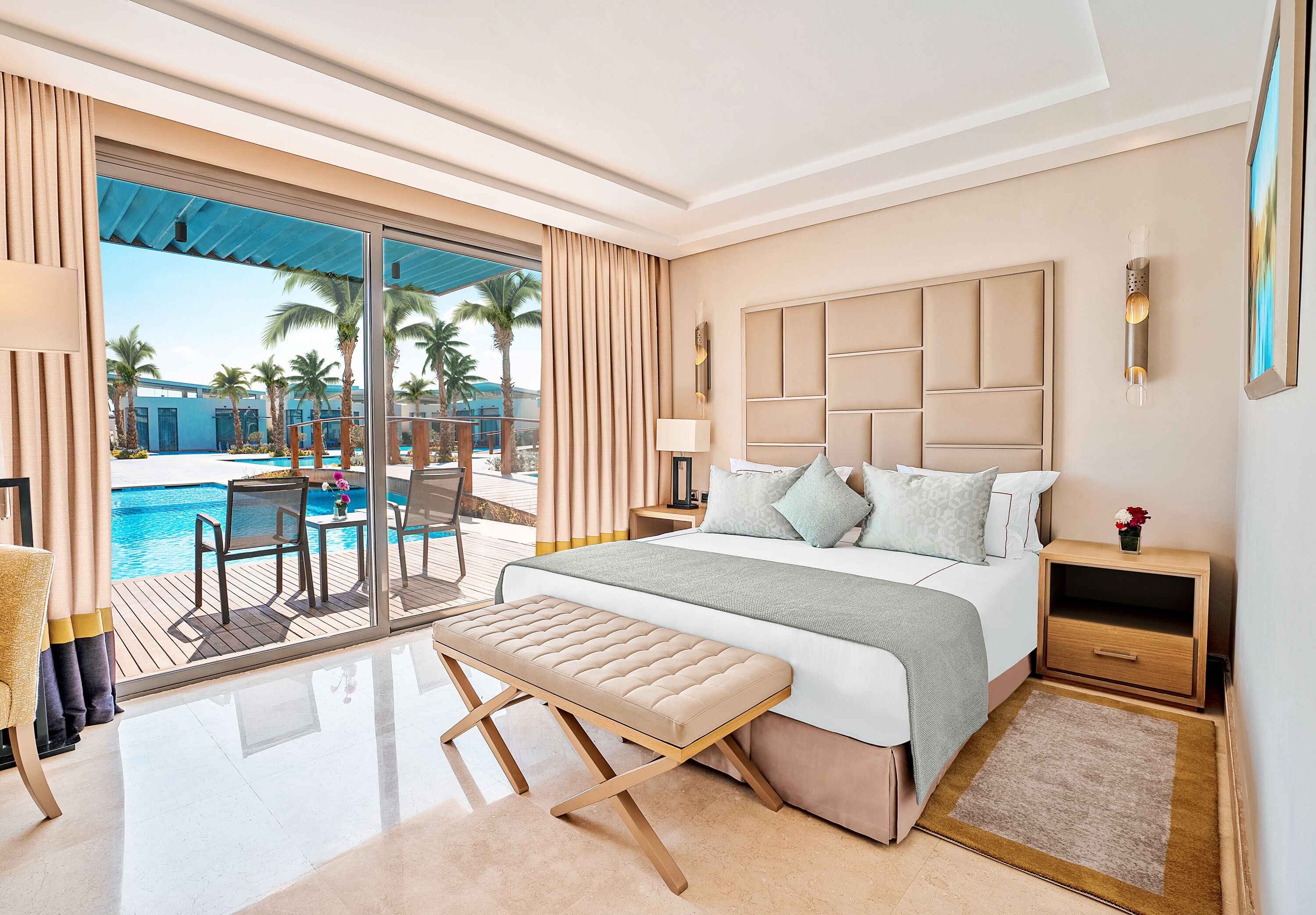 A modern hotel room with a large bed, a bench, a nightstand, a lamp, and a view of a pool and palm trees.