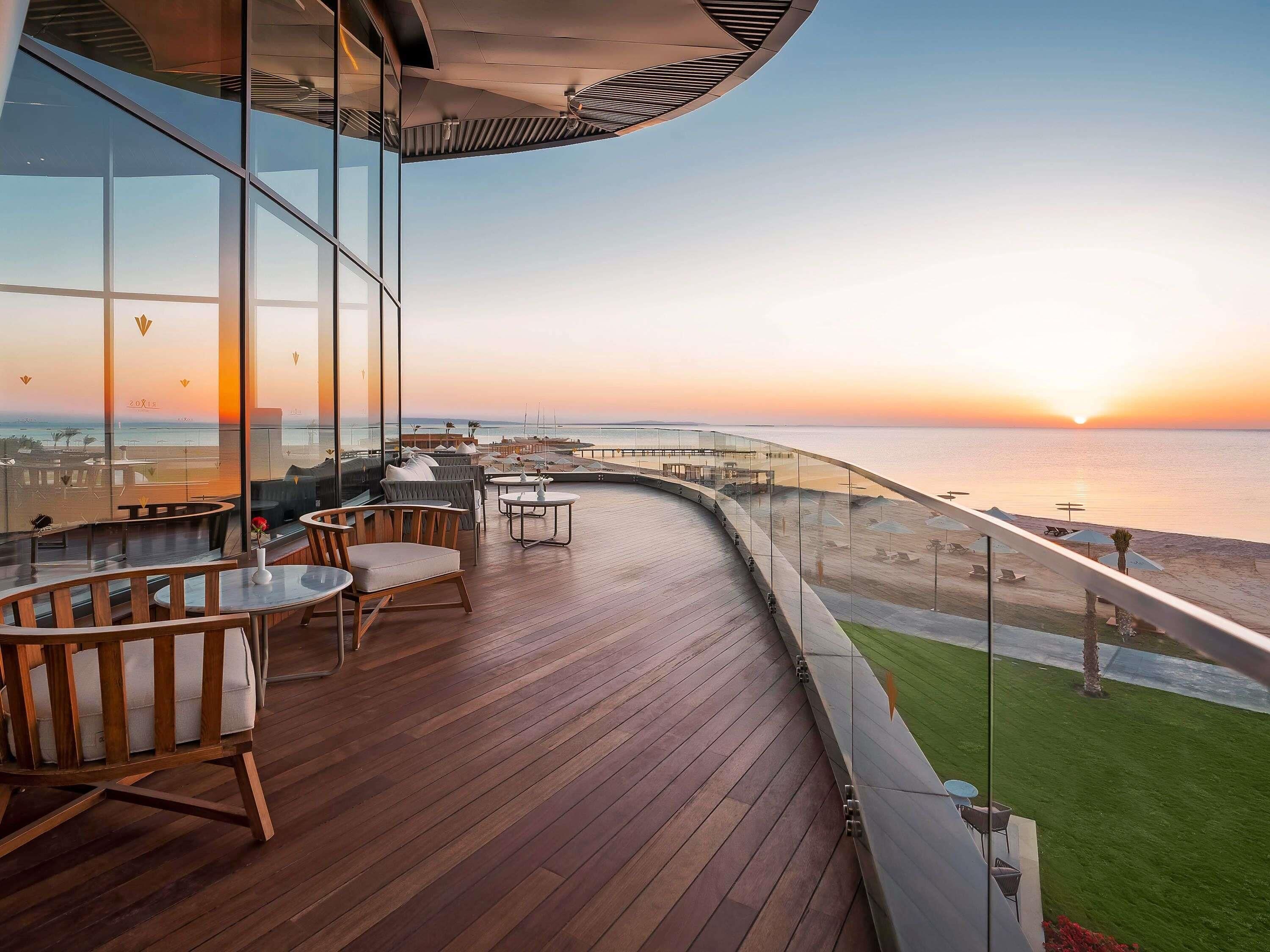 A deck with wooden flooring and seating, overlooking a beach and ocean at sunset.