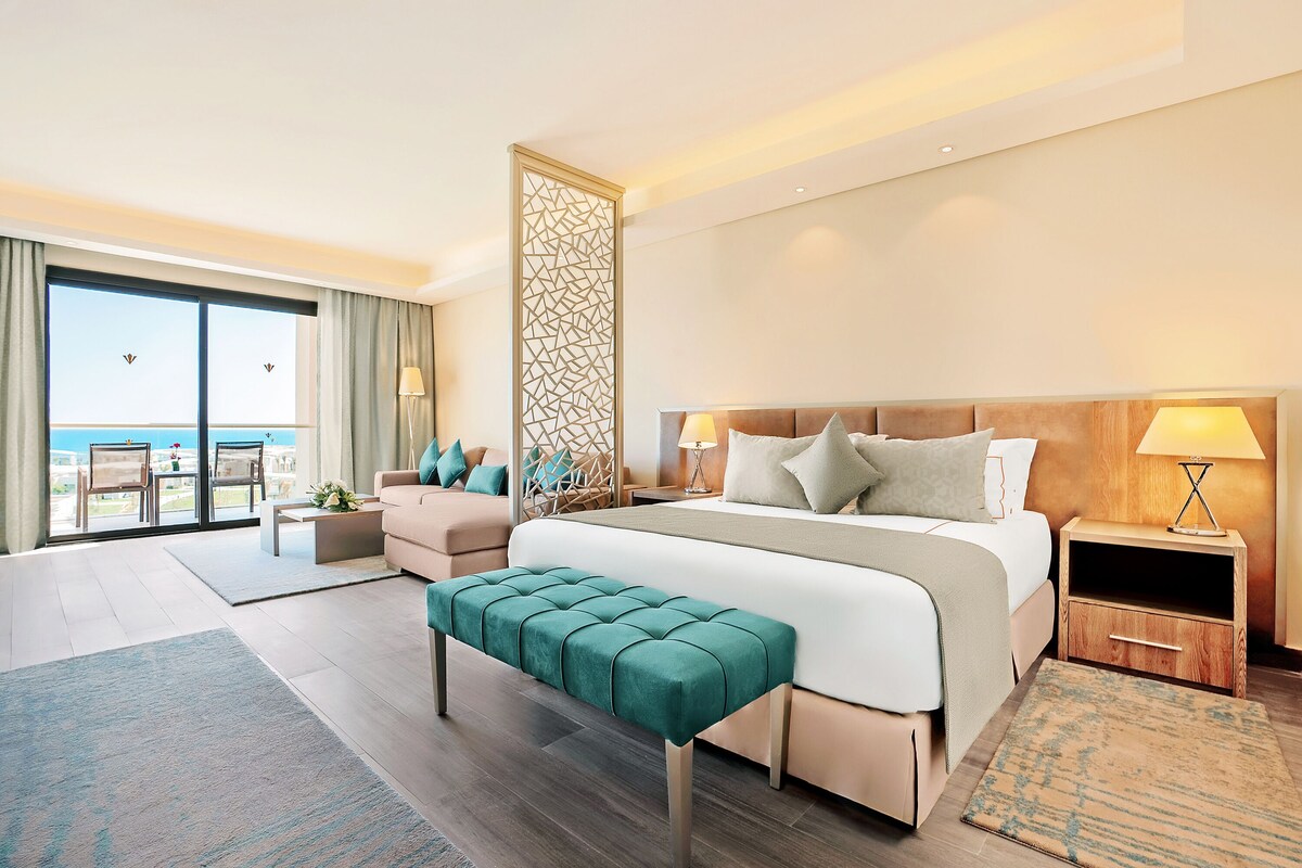 Une chambre d’hôtel moderne avec un grand lit, une banquette, une table de chevet et une vue sur l’océan.
