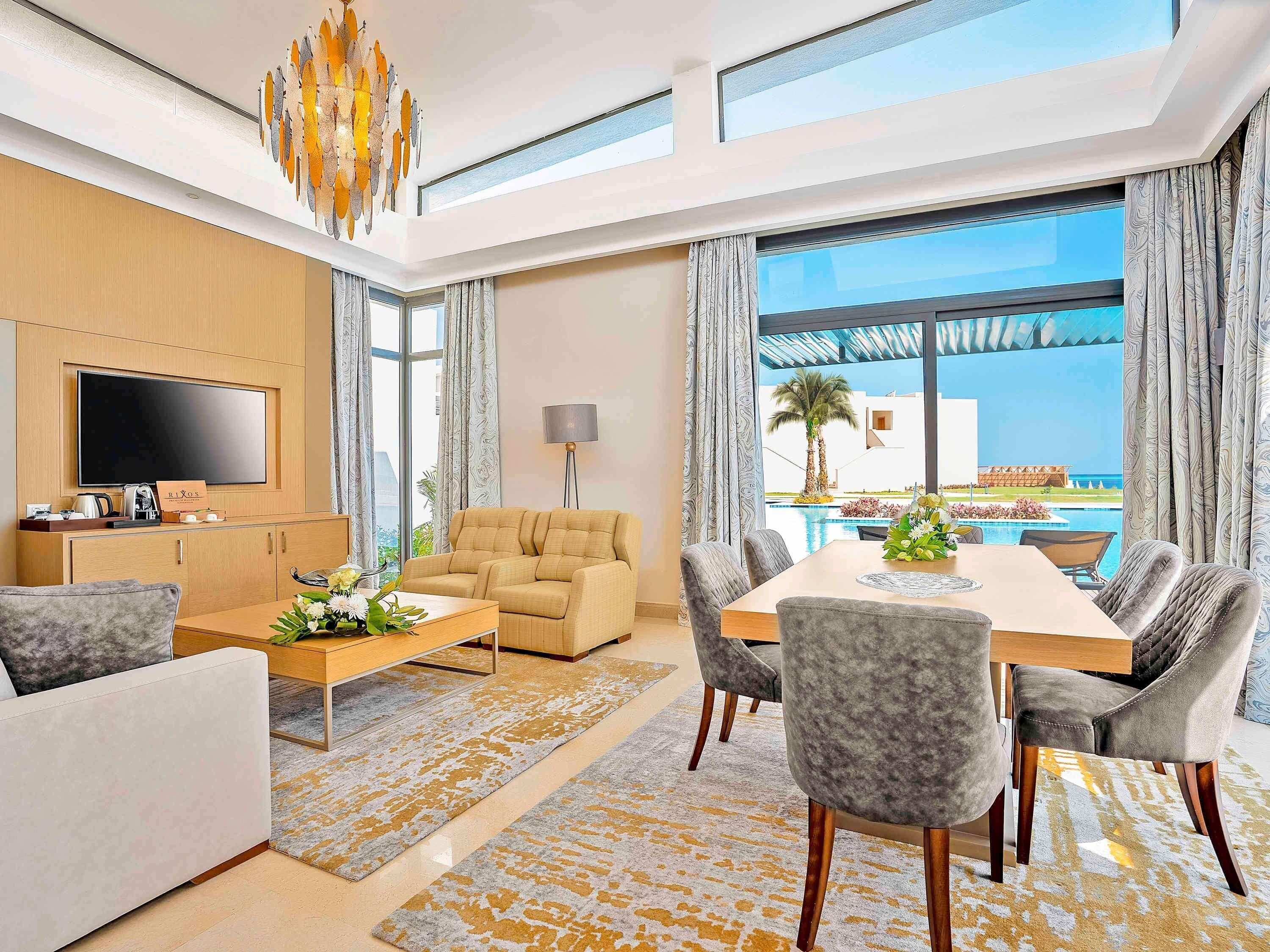 A modern hotel room with a flat-screen TV, a sofa, a dining table with chairs, a coffee station, and a large window with a view of the pool and palm trees.