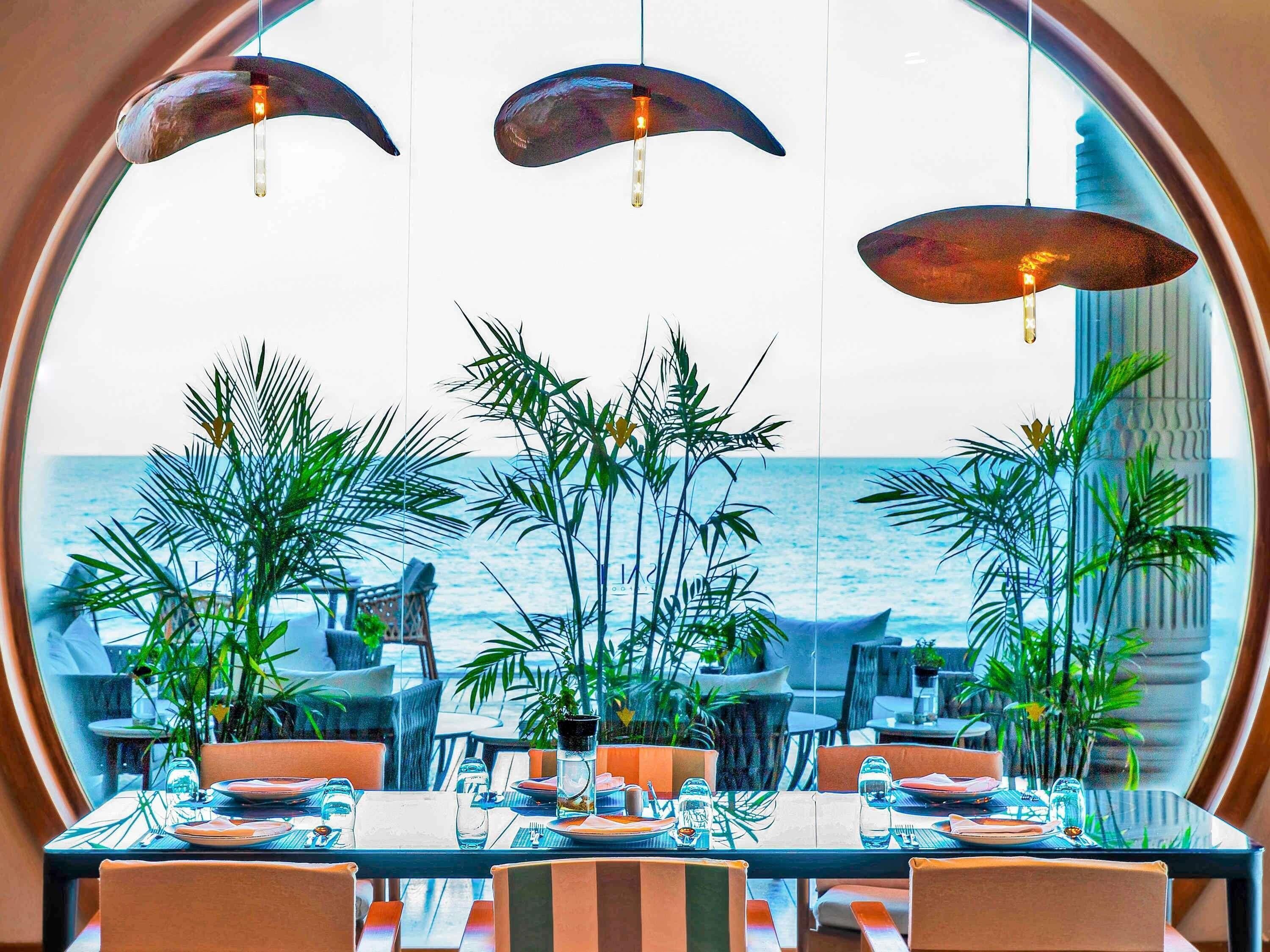A dining area with tables set for four, large windows with a view of the ocean, and hanging pendant lights.