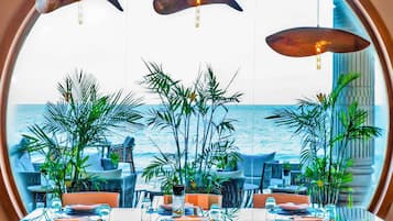 A dining area with tables set for four, large windows with a view of the ocean, and hanging pendant lights.