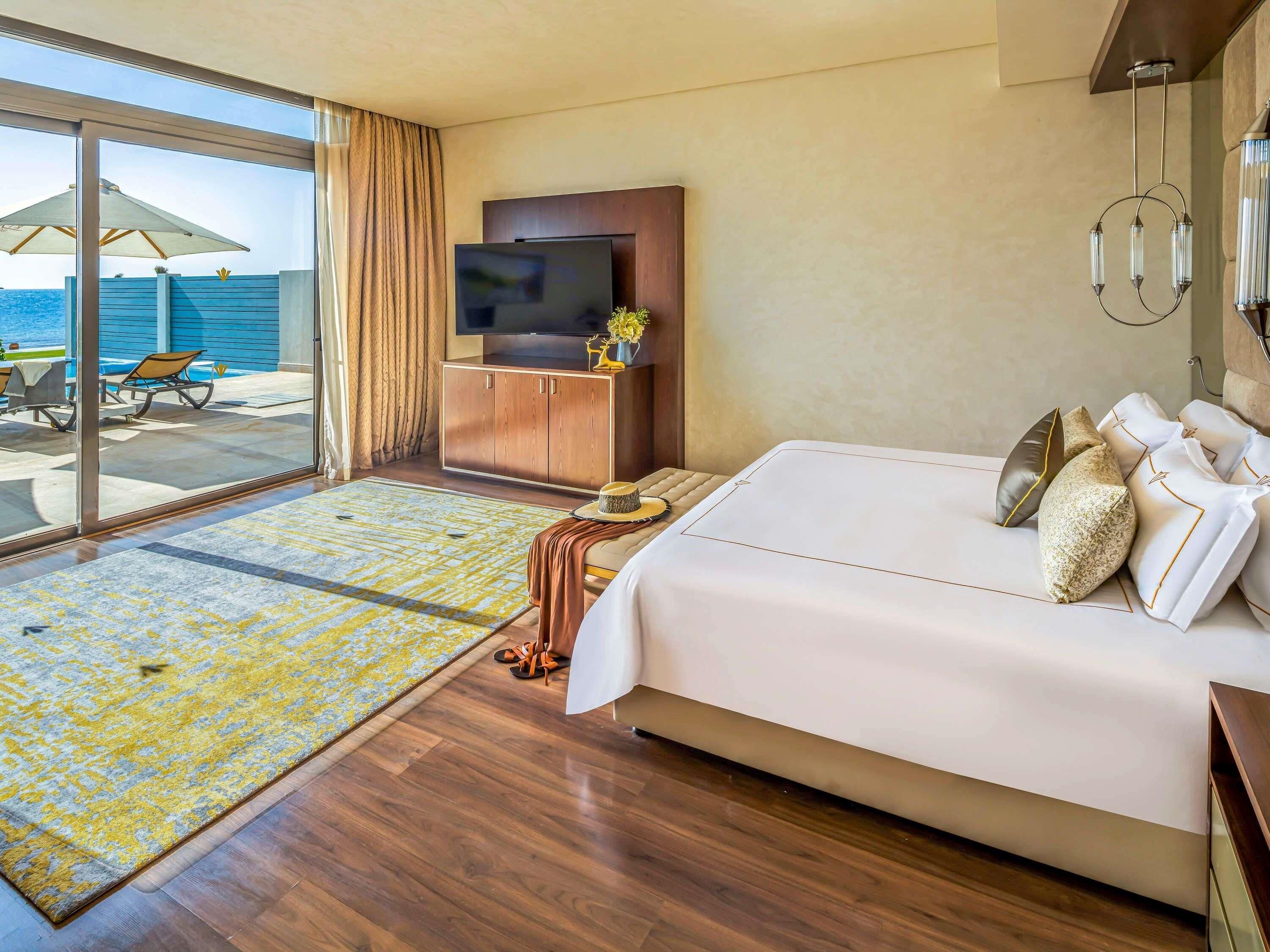A hotel room with a bed, a TV, a wooden cabinet, a large window with curtains, and a view of the sea.