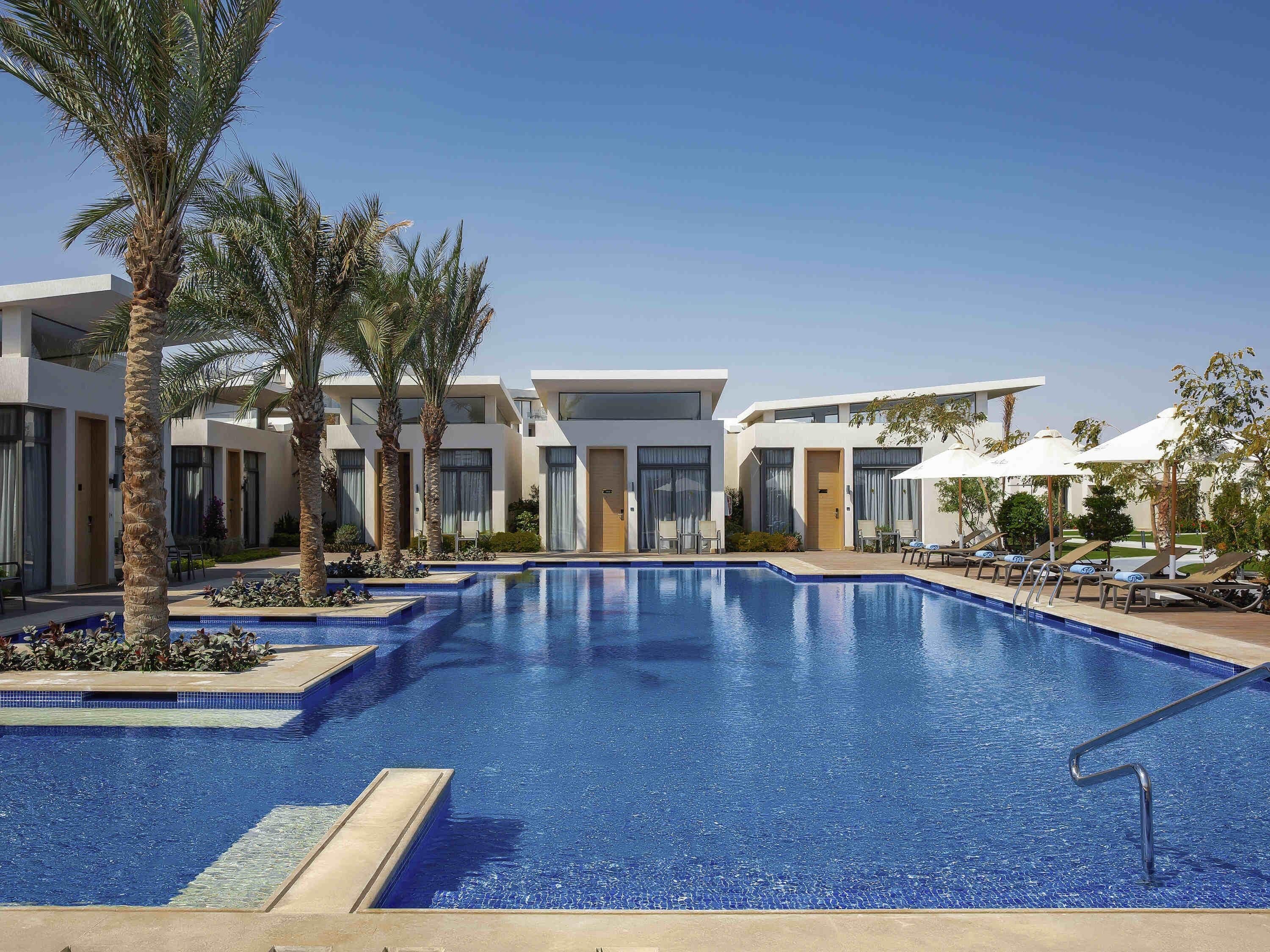 A modern hotel with a central swimming pool, palm trees, and sun loungers.