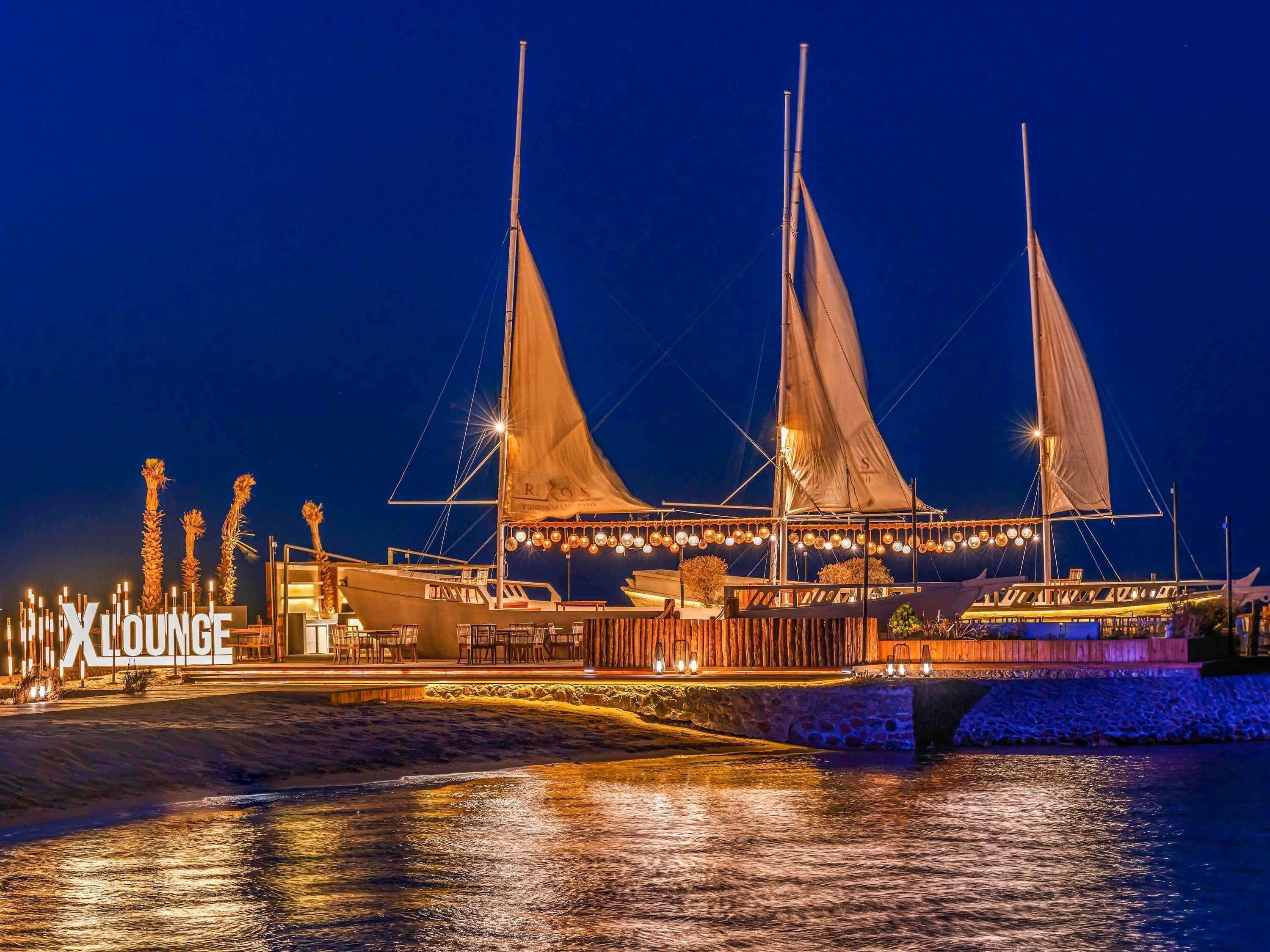 A waterfront venue with a large sailboat, a bar area, and a sign reading 'ALOONGE.'