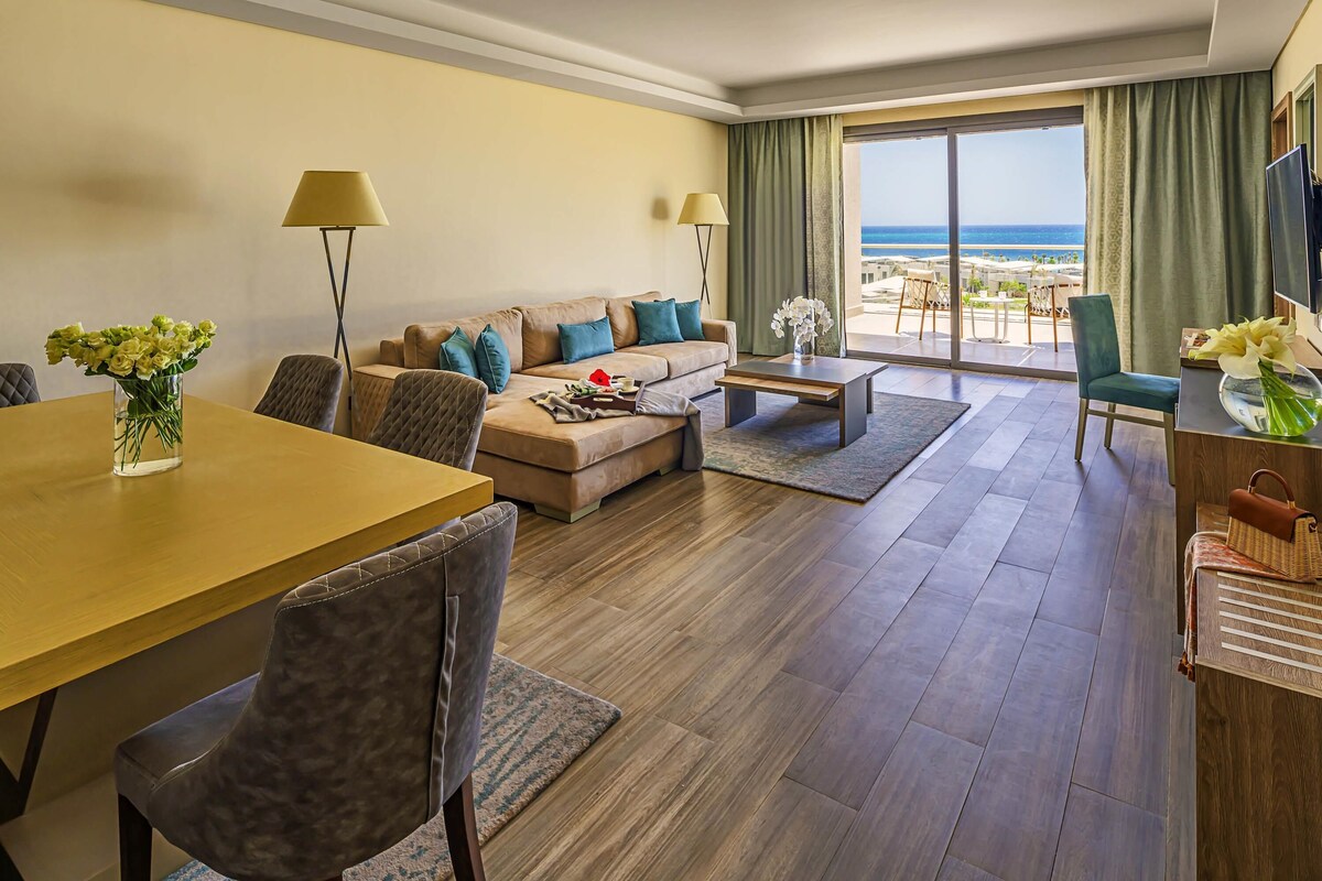Une chambre d’hôtel moderne équipée d’un canapé, d’une table basse, d’une table à manger et de chaises. De grandes fenêtres offrent une vue sur l’océan.