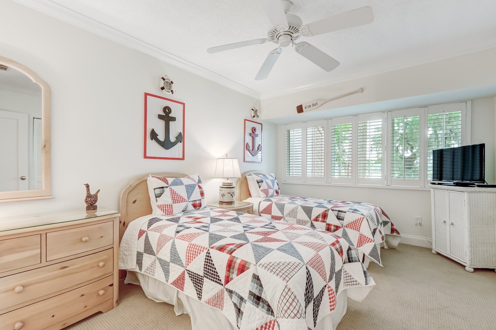 A bedroom with two beds, a dresser, a ceiling fan, a TV, and windows with blinds.