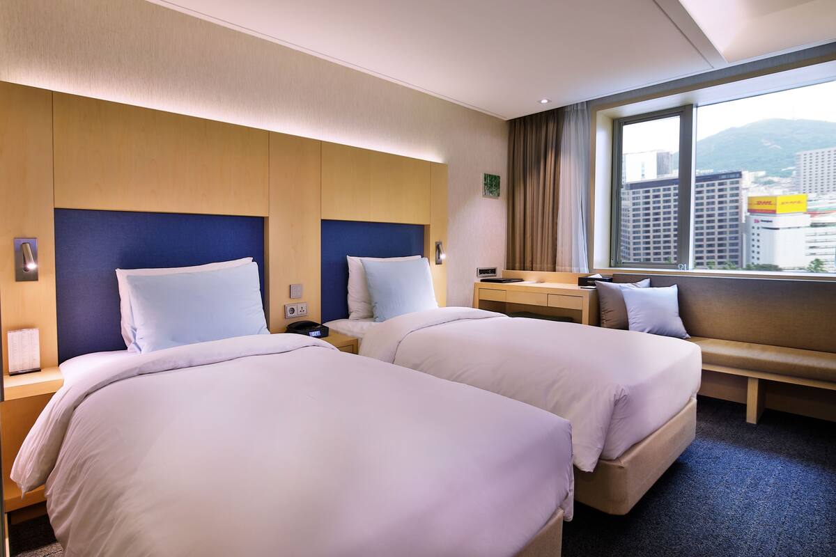 A hotel room with two beds, a desk, and a window with a city view.