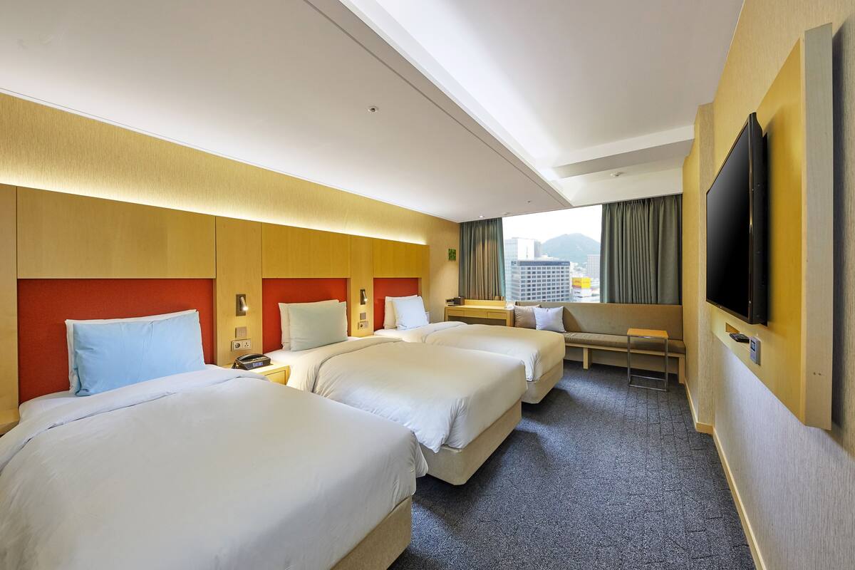 A hotel room with three beds, a TV, a desk, and a window with curtains.