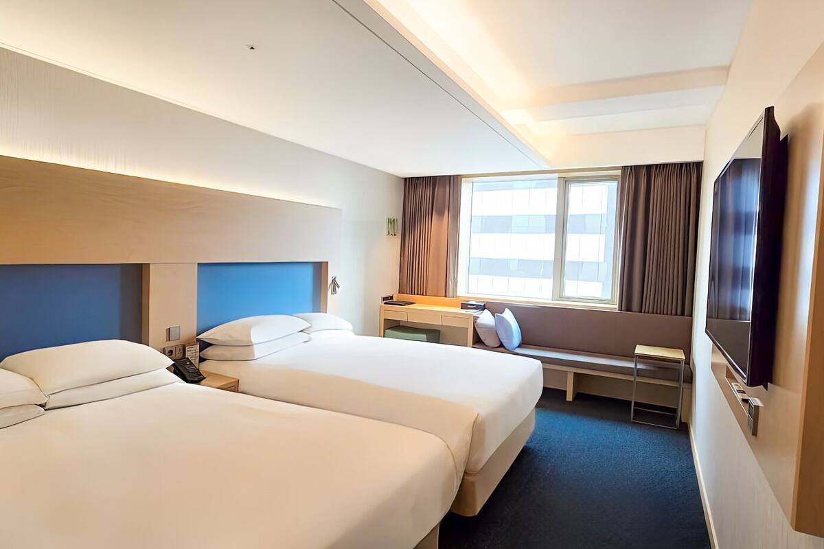 A hotel room with two beds, a desk, a TV, and a window with curtains.