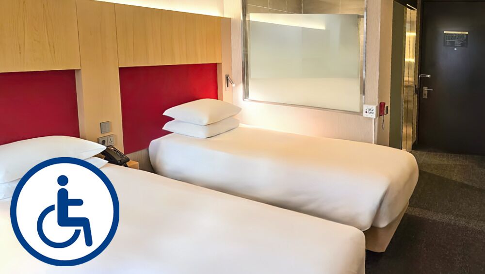 Quarto de hotel com duas camas de solteiro, uma parede de destaque vermelha, cabine de chuveiro em vidro e símbolo de acessibilidade para cadeiras de rodas.