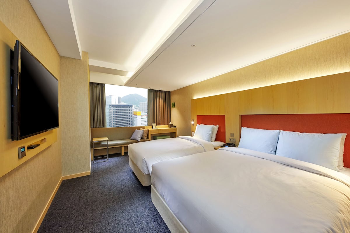 A hotel room with two beds, a desk, a TV, and a view of the city.