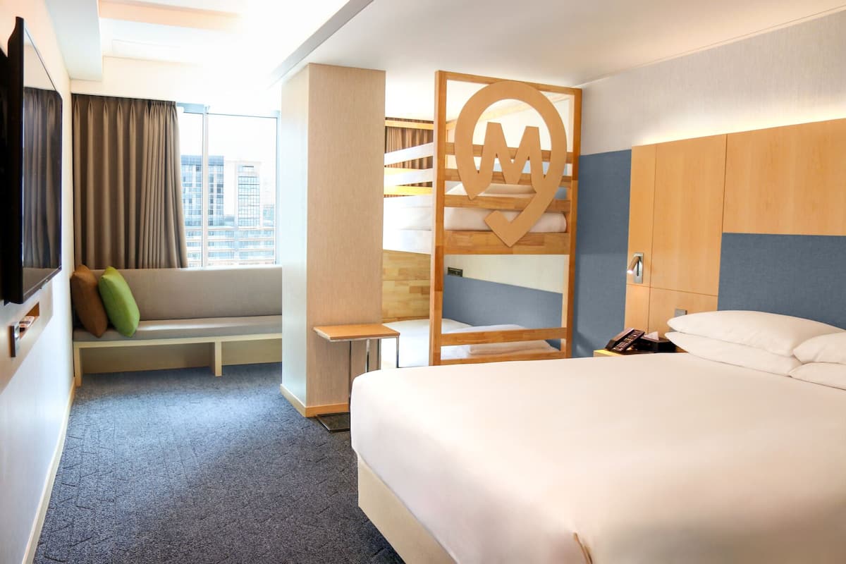 A hotel room with a bunk bed, a desk, a chair, a TV, and a large window with curtains.