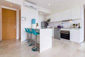 A modern kitchen with white cabinets, a stainless steel refrigerator, a microwave, and a dishwasher. There are two blue bar stools at a central island. - Beautiful 2 BDR condo in Downtown (Playa del Carmen)