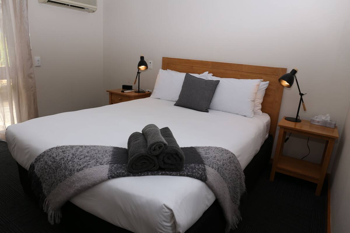 A neatly made bed with white linens and a grey blanket, two bedside lamps, and a wooden headboard.