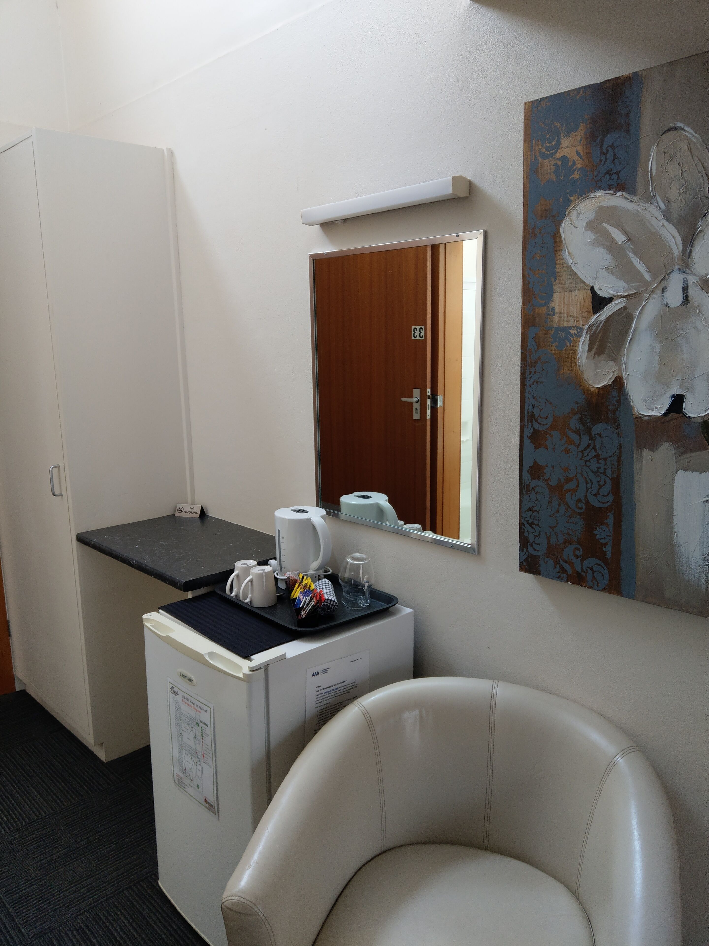 A hotel room with a desk, a minibar, a chair, a painting, and a door.