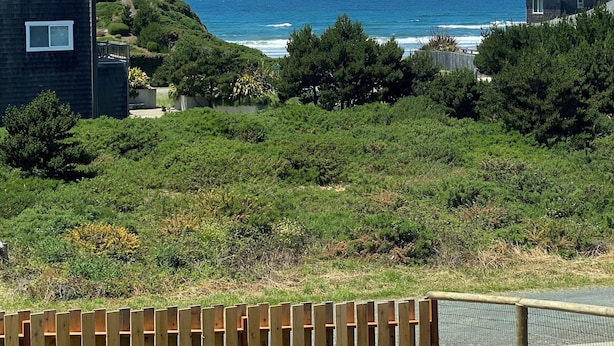 A coastal view with a wooden fence, a small building, and a clear blue sky. - Ocean view, 100 yards to beach, pet friendly, secure fenced yard. (Bandon)