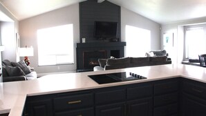 A modern kitchen with a large island, dark cabinetry, a flat-screen TV above the fireplace, and a cozy seating area with a sofa and armchairs. - Ocean view, 100 yards to beach, pet friendly, secure fenced yard. (Bandon)