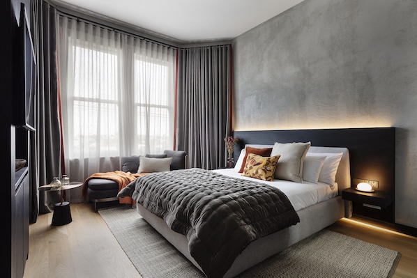 A modern bedroom with a large bed, bedside tables, a TV, and a view of the outdoors through sheer curtains.