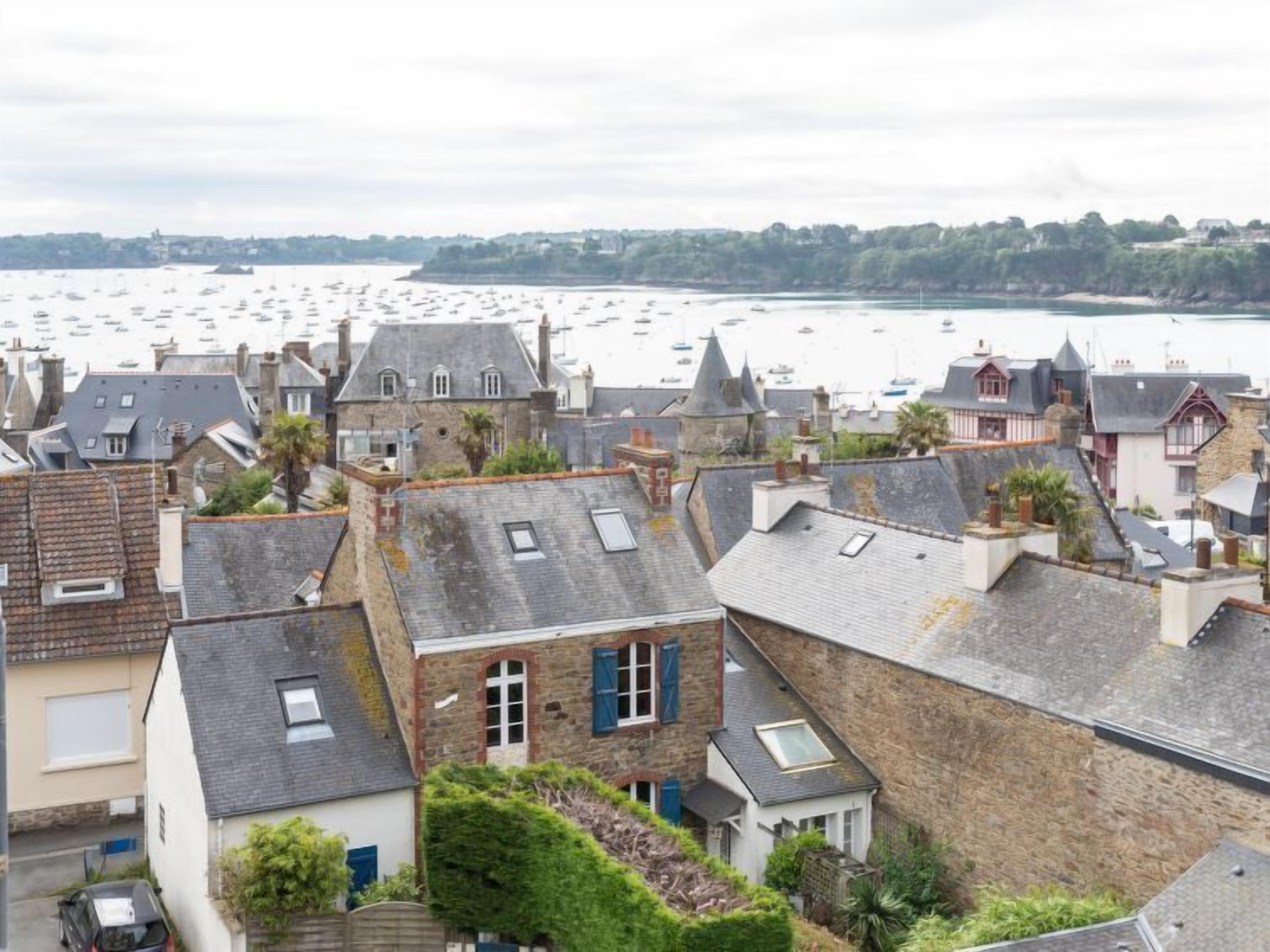 A coastal town with stone buildings, some with sloped roofs and dormer windows, and a view of the sea with numerous boats.