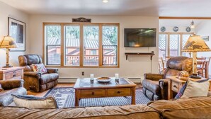 A cozy living room with leather sofas, a wooden coffee table, and a large window with a view of a wooden deck. - Aspen Meadows #2 Sun-Filled, 3 bedroom condo (June Lake)
