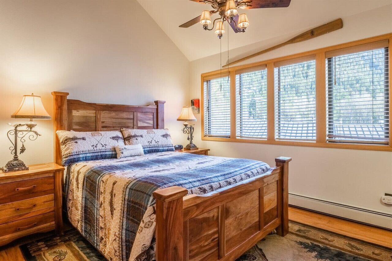 Une chambre à coucher avec un lit en bois, deux tables de chevet avec des lampes, un ventilateur de plafond et une grande fenêtre avec des stores.