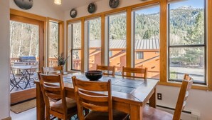 A dining area with a wooden table and chairs, large windows with a mountain view, and a barbecue grill outside. - Aspen Meadows #2 Sun-Filled, 3 bedroom condo (June Lake)