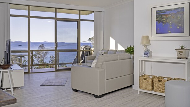 A modern living room with a large window overlooking a body of water, a white sofa, a small table, and a framed painting on the wall. - Nobby's 9 - Amazing Ocean Views (Port Macquarie)