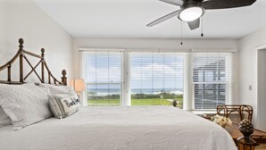 A bedroom with a large bed, a ceiling fan, and a view of the ocean through the windows.