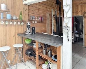 Une cuisine compacte aux murs en bois, avec une table centrale et des tabourets. Elle comprend une machine à café, des étagères remplies d’ustensiles de cuisine et un attrape-rêves.