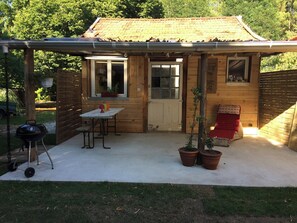 Une cabane en bois avec un toit en tuiles, une petite terrasse avec une table et des chaises, un barbecue et des plantes en pot.