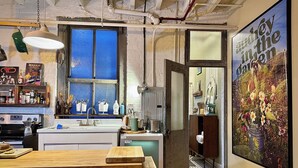 A kitchen with a wooden counter, a sink, and a window with blue blinds. There is a poster on the wall and a door leading to another room.