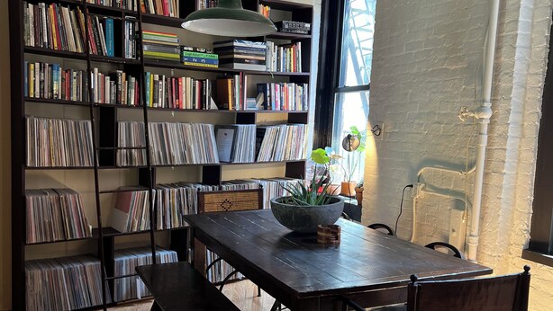 A room with a wooden table, benches, a bookshelf filled with books and CDs, a hanging light fixture, and a patterned rug.