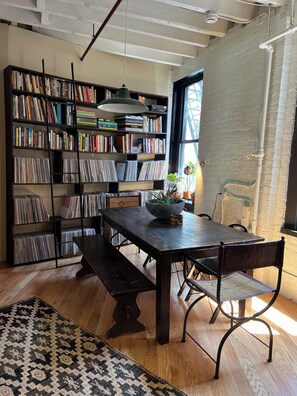 A room with a wooden table, benches, a bookshelf filled with books and CDs, a hanging light fixture, and a patterned rug.