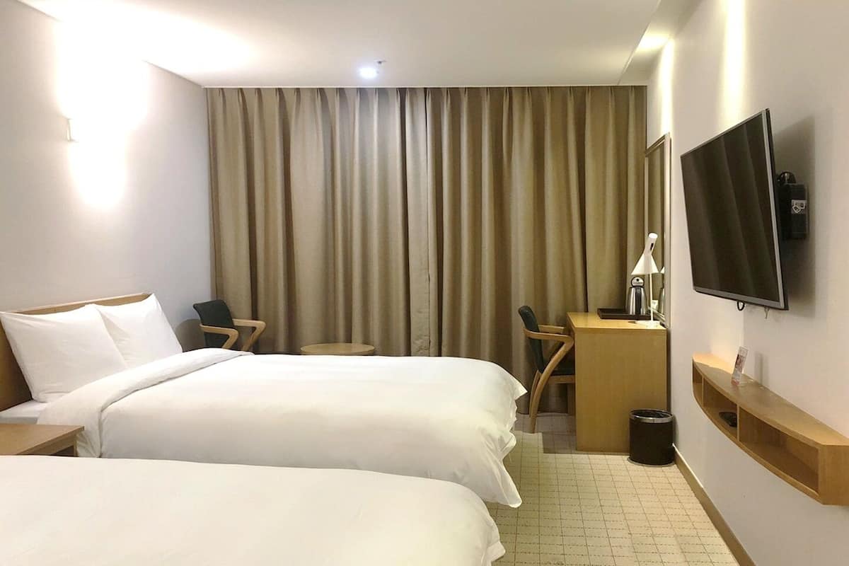 A hotel room with two beds, a desk, a chair, a TV, and a trash bin.