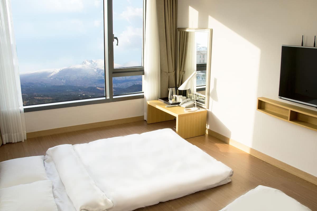 A hotel room with a large window, a bed, a desk with a lamp, a telephone, and a flat-screen TV mounted on the wall.