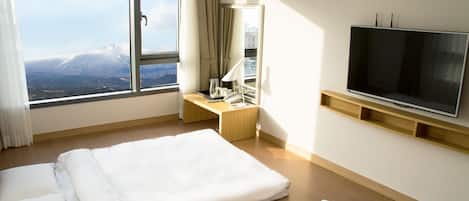 A hotel room with a large window, a bed, a desk with a lamp, a telephone, and a flat-screen TV mounted on the wall.